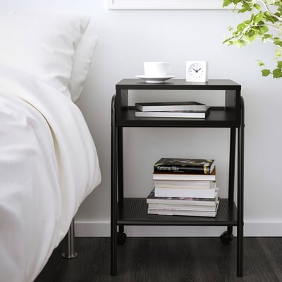 Kleiner Nachttisch aus schwarzem Metall neben einem Bett mit Büchern, weißer Schüssel und Wecker.