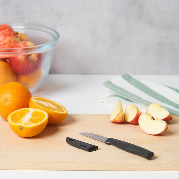 Ein Schneidebrett aus Holz mit einem Gemüsemesser, Orangenhälften, Apfelscheiben und einer Schüssel mit Äpfeln. Das Messer hat einen schwarzen Griff und eine schwarze Klingenabdeckung.