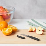Ein Schneidebrett aus Holz mit einem Gemüsemesser, Orangenhälften, Apfelscheiben und einer Schüssel mit Äpfeln. Das Messer hat einen schwarzen Griff und eine schwarze Klingenabdeckung.