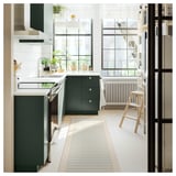Moderne Küche mit grünen Schränken und einem gestreiften blau-weißen Teppich. Großes Fenster, Topfpflanzen und Holzhocker.