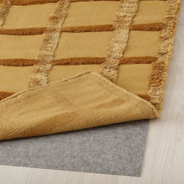 Gelb gemusterter Teppich mit erhabenem Gittermuster, verlegt auf hellem Holzboden, graue Filzunterlage an der Ecke sichtbar.