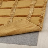 Gelb gemusterter Teppich mit erhabenem Gittermuster, verlegt auf hellem Holzboden, graue Filzunterlage an der Ecke sichtbar.
