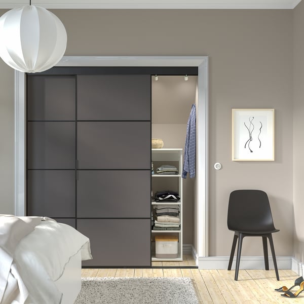 Modernes Schlafzimmer mit grauem SKITTA-Kleiderschrank, gepflegter Kleidung, schwarzem Stuhl, weißem Licht, Bett und Wandkunst.