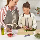 Frau und Kind bereiten mit Schürzen Essen zu. Das Kind benutzt ein kindersicheres Messer, um Gurken zu schneiden.