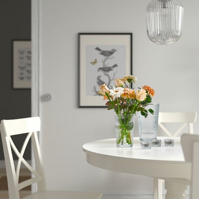 Kleine Glasvase mit Blumen auf einem weißen Tisch, neben hohem Glas.