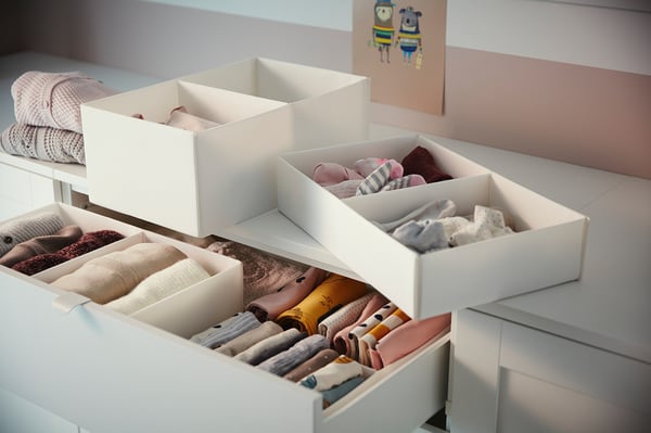 Weiße SMÅSTAD Kinderkommode mit vier offenen Schubladen, die organisierte Babykleidung in weißen und beigen Farben zeigt.