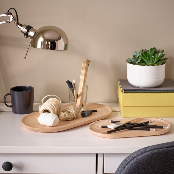 Schreibtisch mit SMÖRLING Tabletts, einem Becher und einer Pflanze. Tabletts bieten Platz für Malutensilien und Seile. Heller und organisierter Arbeitsbereich mit einer Lampe.