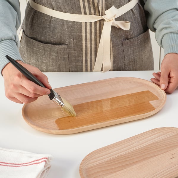 Die Person bestreicht eine orangefarbene Flüssigkeit auf ein ovales Serviertablett aus Holz. Das Tablett wird auf einen weißen Tisch gestellt, bereit zum Servieren von Speisen oder zur Dekoration.