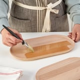 Die Person bestreicht eine orangefarbene Flüssigkeit auf ein ovales Serviertablett aus Holz. Das Tablett wird auf einen weißen Tisch gestellt, bereit zum Servieren von Speisen oder zur Dekoration.