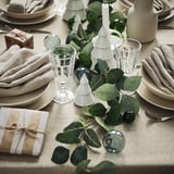 Weihnachtstisch gedeckt mit weißen Tellern, Gläsern, grauen Servietten und Geschenken. In der Mitte befinden sich SMYCKA-Girlanden mit Plastikblättern. Kleine beleuchtete Bäume schmücken.