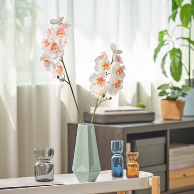 Weiße Orchidee in grüner geometrischer Vase, kleine blau/orangefarbene Vasen daneben, in der Nähe des Fensters mit Pflanze.