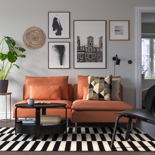 Modernes Wohnzimmer mit orangefarbener SÖDERHAMN-Couch, schwarz-weißem Teppich, Kunstgaleriewand, schwarzem Couchtisch und Pflanze.