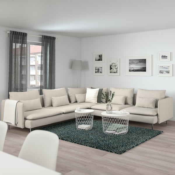 Ein beiges Ecksofa von SÖDERHAMN mit grünem Teppich, Beistelltischen und Kissen in der Nähe eines großen Fensters mit Vorhängen, das Komfort und Größe betont.