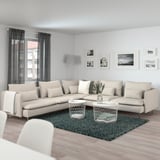 Ein beiges Ecksofa von SÖDERHAMN mit grünem Teppich, Beistelltischen und Kissen in der Nähe eines großen Fensters mit Vorhängen, das Komfort und Größe betont.