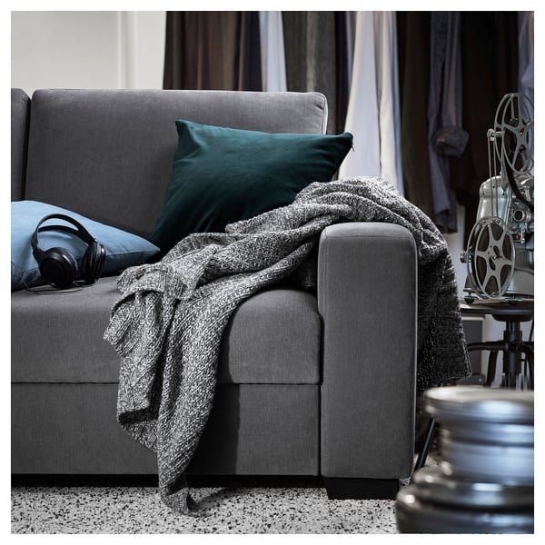Ein modernes graues SÖRVALLEN-Sofa mit einem grünen Kissen und einer gemütlichen grauen Decke. Darauf ruhen Kopfhörer mit einer Filmrolle in der Nähe.