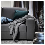 Ein modernes graues SÖRVALLEN-Sofa mit einem grünen Kissen und einer gemütlichen grauen Decke. Darauf ruhen Kopfhörer mit einer Filmrolle in der Nähe.