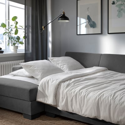 Modernes graues Schlafsofa mit weißer Bettwäsche, Wandlampe und Pflanze in der Nähe des Fensters.