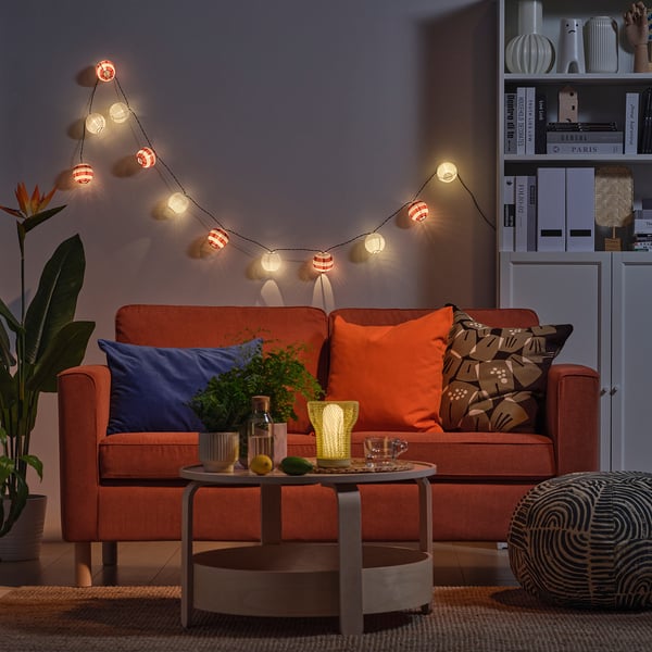 Gemütliches Wohnzimmer mit orangefarbenem Sofa, Kissen, Lichterketten und Couchtisch mit Zitrusfrüchten, Mondlampe und Pflanze.