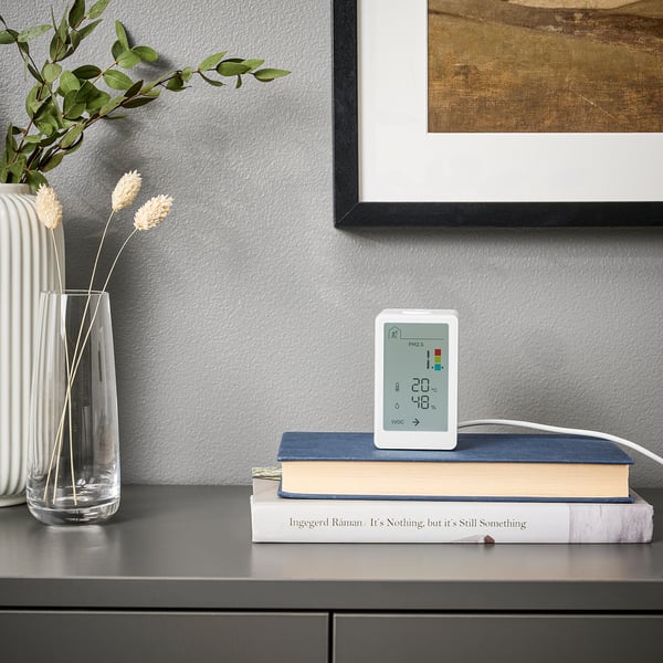 Ein eleganter weißer Luftqualitätsmonitor, der auf Büchern in der Nähe von von der Natur inspirierten Dekorationen angebracht ist. Es zeigt Temperatur, Luftfeuchtigkeit, pm2 an. 5 und VOC-Pegel.