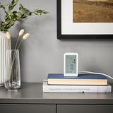 Ein eleganter weißer Luftqualitätsmonitor, der auf Büchern in der Nähe von von der Natur inspirierten Dekorationen angebracht ist. Es zeigt Temperatur, Luftfeuchtigkeit, pm2 an. 5 und VOC-Pegel.
