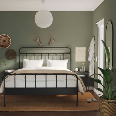 Grünes Schlafzimmer mit schwarzem Bett, weißer Bettwäsche, gewebter Wanddekoration, Hängelampen und einer hohen Pflanze.