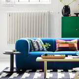 Modernes Wohnzimmer mit blauem STOCKHOLM 2025 Sofa, lebendigen Kissen, Couchtisch, geometrischem Teppich, grünem Schrank und großen Fenstern.