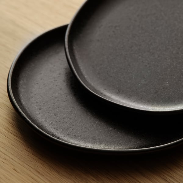 Zwei mattschwarze Teller mit dezenten Sprenkeln, ordentlich gestapelt auf einem Holztisch. Rustikale, handwerkliche Optik.