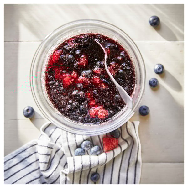 Ein zylindrisches Glasgefäß, gefüllt mit dunkler Beerenmarmelade, mit einem Löffel und umgeben von verstreuten Heidelbeeren.
