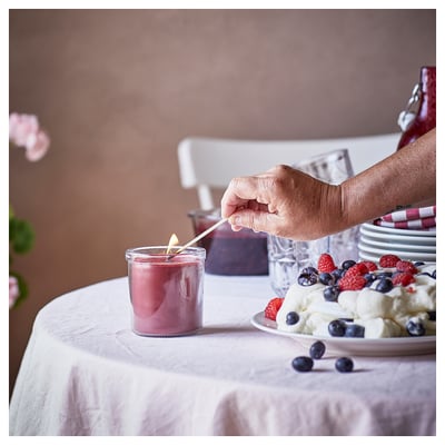 Person zündet rosa Kerze in der Nähe von Beerendessert an. Tisch gedeckt mit weißem Tuch, Tellern, Blumen und Flasche. Warme, einladende Atmosphäre.