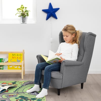 Kind liest im grauen STRANDMON Sessel. Heller Teppich, Bücherregal mit Büchern daneben. Blauer Stern Wanddekoration, Pflanze am Fenster.