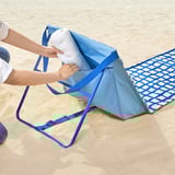 Eine Person legt ein weißes Handtuch in eine blaue Stofftasche mit Reißverschlüssen von STRANDÖN Sonnenliegen an einem Sandstrand.