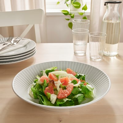 Eine Schüssel mit Grapefruit-Salat steht auf einem hellen Holztisch am Fenster, umgeben von leeren Gläsern und gestapelten Tellern mit Besteck.