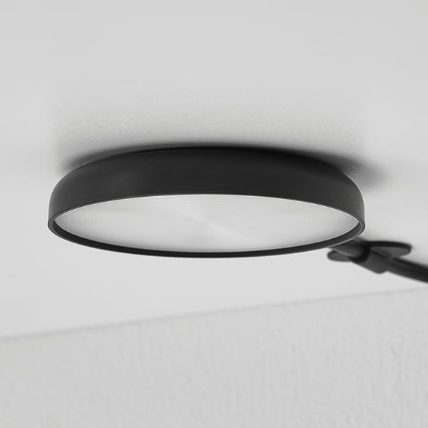 Moderne schwarze Deckenleuchte teilweise sichtbar, montiert an einer weißen Wand. Strahlt ein helles, gleichmäßiges Leuchten aus.