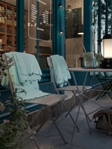 Zwei blaue SUNDSÖ-Klappstühle in der Nähe eines Café-Eingangs mit mintfarbenen Decken und Gläsern auf dem Tisch.