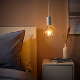 Gemütliche Schlafzimmerecke mit SUNNEBY-Lampe, orangefarbener Glühbirne, blauem und gelbem Kissen, Nachttisch mit Buch und Vase, warmer brauner Wand und moderner Malerei.
