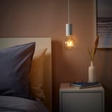 Gemütliches Schlafzimmer mit SUNNEBY Pendelleuchte über dem Nachttisch, warmem Licht, Büchern, Trockenblumen und blau-gelben Kissen.