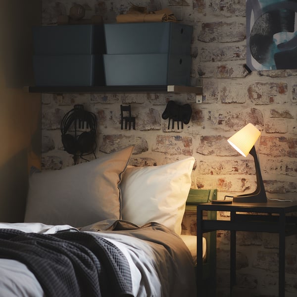 Gemütliche Schlafzimmerecke mit freiliegender Ziegelwand, weißen Kissen, grauer Decke, kleinem schwarzen Tisch und moderner schwarzer Lampe.