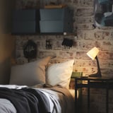 Gemütliche Schlafzimmerecke mit freiliegender Ziegelwand, weißen Kissen, grauer Decke, kleinem schwarzen Tisch und moderner schwarzer Lampe.