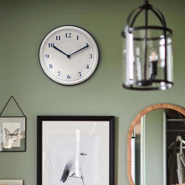 Eine runde Wanduhr mit weißem, emailleartigem Zifferblatt und blauen Ziffern. Es zeigt die Zeit an, ohne zu ticken. Hängt über einem gerahmten Naturkunstwerk an einer grünen Wand.