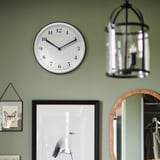 Eine runde Wanduhr mit weißem, emailleartigem Zifferblatt und blauen Ziffern. Es zeigt die Zeit an, ohne zu ticken. Hängt über einem gerahmten Naturkunstwerk an einer grünen Wand.