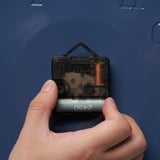 Hand mit einer kleinen, quadratischen Uhr mit transparenter Abdeckung, die den Blick auf die internen Mechanismen freigibt.