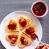 Herzförmige Waffeln mit roter Marmelade und Erdbeeren auf einem weißen Teller. Daneben einen Löffel mit Marmelade.