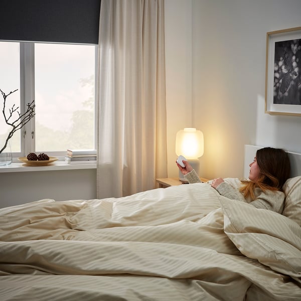 Eine Frau liegt im Bett und hält eine Fernbedienung in der Nähe einer modernen Lampe, die die Verwendung der SYMFONISK Smart-Home-Fernbedienung veranschaulicht.