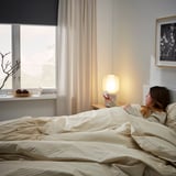 Eine Frau liegt im Bett und hält eine Fernbedienung in der Nähe einer modernen Lampe, die die Verwendung der SYMFONISK Smart-Home-Fernbedienung veranschaulicht.