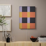 Eine bunte geometrische Kunsttafel hängt an der Wand, Teil des SYMFONISK WLAN-Lautsprechers, neben Dekorationsgegenständen.