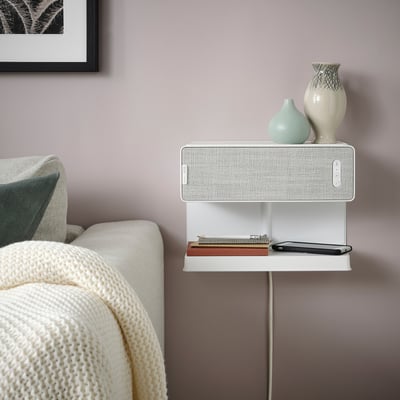 SYMFONISK weißer Regallautsprecher mit kabellosem Ladegerät, an der Wand. In der Nähe eines gemütlichen Sofas mit Decke, Büchern und Dekoration.