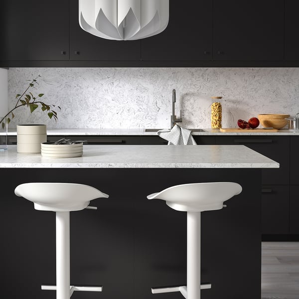 Moderne Küche mit schwarzen Schränken, weißer Theke, Hockern, Pendelleuchte, Pflanze, Schalen, goldener Vase, Geschirrkorb, weißer Rückwand.