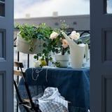 Balkonszene: helle Holzkübel mit rosa Blumen, weiße Gießkanne, blaue Tischdecke, schwarzer Metallstuhl mit karierter Decke.