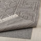 Grauer, handgewebter Wollteppich mit geometrischem Muster liegt auf hellem Holzboden. Erhabene Muster und Fransendetails sind sichtbar.