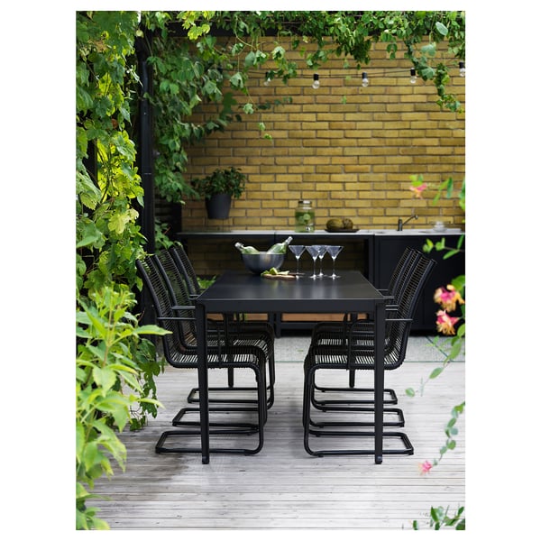 Modernes Esszimmer im Freien mit schwarzen Stühlen und Tisch auf einer Holzterrasse mit gelber Backsteinmauer im Hintergrund und üppigem Grün.